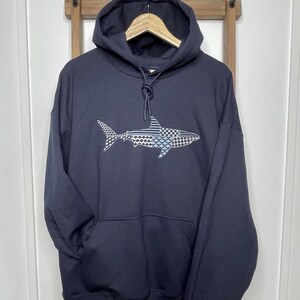 Embroidered navy blue hooded sweatshirt with a great white shark design. The shark is composed of various geometric patterns in shades of blue, gray, and white. The sweatshirt has a front kangaroo pocket and a drawstring hoodie. The 12 inch design is centered on the front of the sweatshirt.