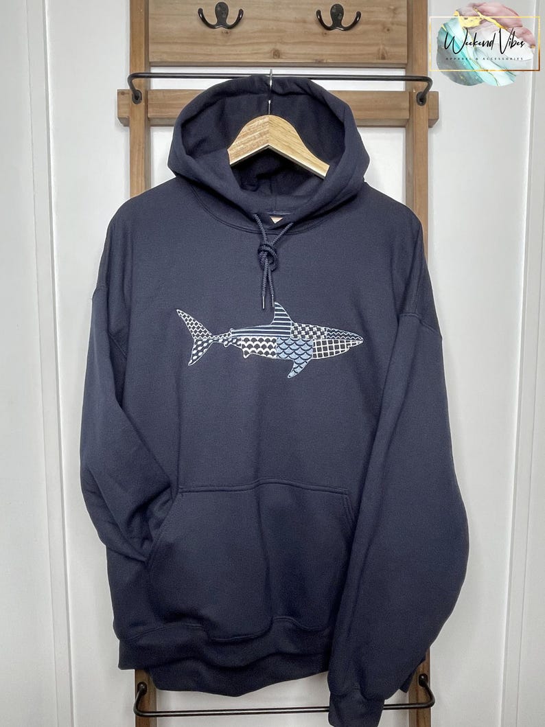 Embroidered navy blue hooded sweatshirt with a great white shark design. The shark is composed of various geometric patterns in shades of blue, gray, and white. The sweatshirt has a front kangaroo pocket and a drawstring hoodie. The 12 inch design is centered on the front of the sweatshirt.