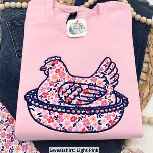 Hen on Nest sweatshirt using a Light Pink sweatshirt, Fabric M, and Navy thread. Photo has fabric swatch, jeans, decorative beads, and a grass basket surrounding sweatshirt.