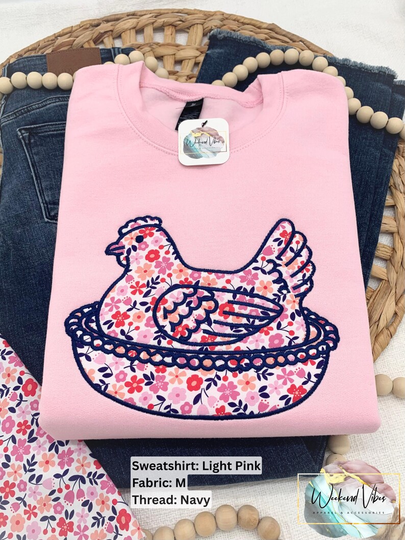 Hen on Nest sweatshirt using a Light Pink sweatshirt, Fabric M, and Navy thread. Photo has fabric swatch, jeans, decorative beads, and a grass basket surrounding sweatshirt.