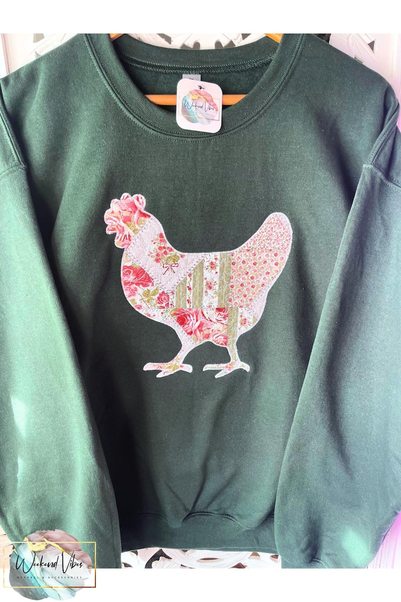 A dark green sweatshirt with an appliqued patchwork floral chicken design. The sweatshirt has a crew neck and long sleeves. The chicken is pink, white, and green with a white zigzag stitch around it. The sweatshirt tag says 'Weekend Vibes Apparel'.