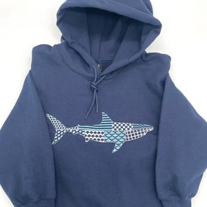 Folded navy blue hooded sweatshirt with a great white shark embroidery design. The shark is made of various blue, gray, and white patterned embroidery thread. The sweatshirt has a drawstring hood and long sleeves. The brand logo "Weekend Vibes" is in the top left corner.