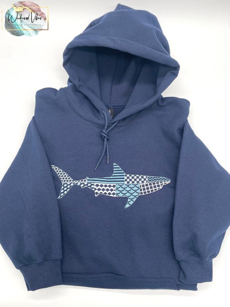 Folded navy blue hooded sweatshirt with a great white shark embroidery design. The shark is made of various blue, gray, and white patterned embroidery thread. The sweatshirt has a drawstring hood and long sleeves. The brand logo "Weekend Vibes" is in the top left corner.