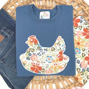 Embroidered Hen on Nest sweatshirt using Indigo Blue sweatshirt and Fabric option D. Photo has a display of the sweatshirt on top of dark jean, the fabric swatch, and a decorative grass basket.