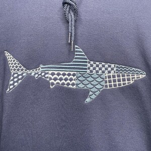 Navy blue hoodie featuring an embroidered great white shark design. The shark is composed of geometric embroidery patterns in shades of blue, gray, and white. The hoodie has a drawstring and a front kangaroo pocket. The text "Weekend Vibes Apparel" is visible at the bottom of photo.