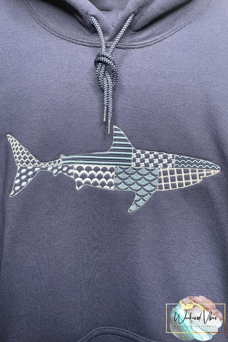 Navy blue hoodie featuring an embroidered great white shark design. The shark is composed of geometric embroidery patterns in shades of blue, gray, and white. The hoodie has a drawstring and a front kangaroo pocket. The text "Weekend Vibes Apparel" is visible at the bottom of photo.