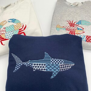 K&ouml;nnte beinhalten: Drei Sweatshirts mit gestickten Meereskreaturen-Designs. Ein marineblaues Sweatshirt zeigt einen Hai, w&auml;hrend ein wei&szlig;es Sweatshirt eine Krabbe hat und ein graues Sweatshirt einen Hummer zeigt. Das Logo "Weekend Vibes" befindet sich oben links.