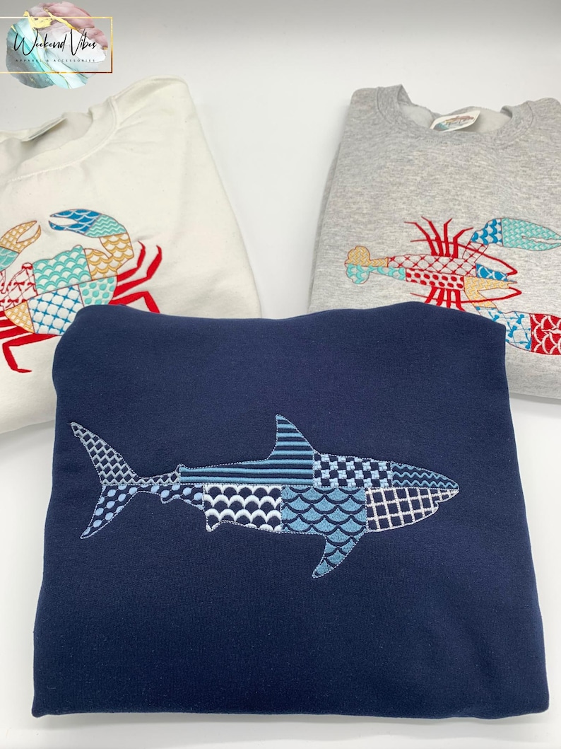 K&ouml;nnte beinhalten: Drei Sweatshirts mit gestickten Meereskreaturen-Designs. Ein marineblaues Sweatshirt zeigt einen Hai, w&auml;hrend ein wei&szlig;es Sweatshirt eine Krabbe hat und ein graues Sweatshirt einen Hummer zeigt. Das Logo "Weekend Vibes" befindet sich oben links.