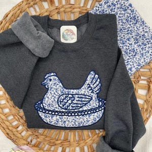 Embroidered Hen on Nest sweatshirt on a Dark Heather Gildan sweatshirt using Fabric B and Navy thread. A swatch of fabric B is in the photo and the sweatshirt is laying on top of a grass basket.