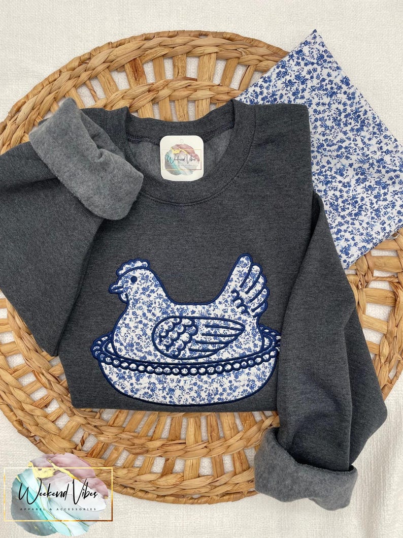 Embroidered Hen on Nest sweatshirt on a Dark Heather Gildan sweatshirt using Fabric B and Navy thread. A swatch of fabric B is in the photo and the sweatshirt is laying on top of a grass basket.