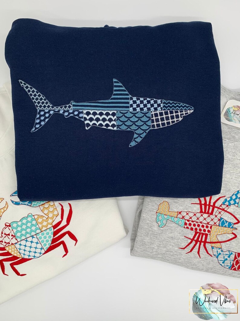 Embroidered navy blue hoodie featuring a great white shark with an embroidery patchwork design in shades of blue, gray, and white. Additional embroidered sweatshirts with a crab and a lobster are visible. The shirts are folded and stacked, showcasing a nautical theme.
