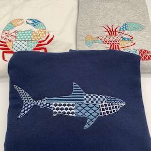 Three sweatshirts with embroidered sea creatures. A white sweatshirt features a crab design with red, teal blue, and gold accents. A gray sweatshirt has a lobster design with red, teal, and gold embroidery. A navy blue sweatshirt has a great white shark design in blues, gray, and white embroidery.