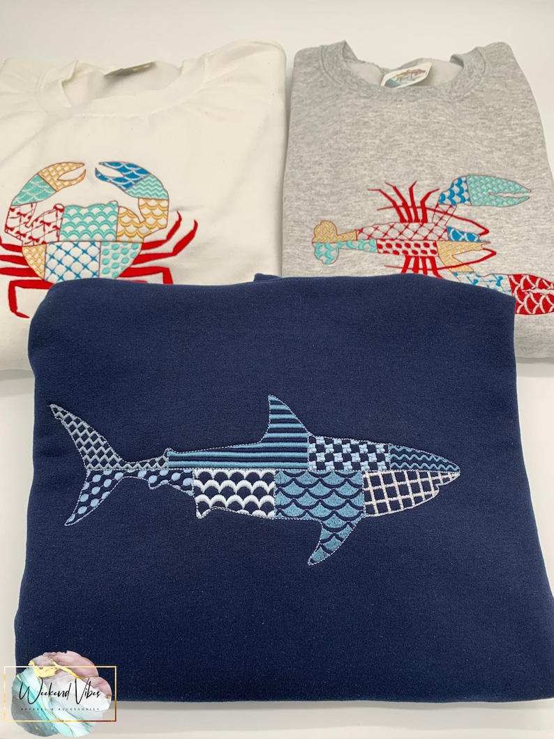 Three sweatshirts with embroidered sea creatures. A white sweatshirt features a crab design with red, teal blue, and gold accents. A gray sweatshirt has a lobster design with red, teal, and gold embroidery. A navy blue sweatshirt has a great white shark design in blues, gray, and white embroidery.
