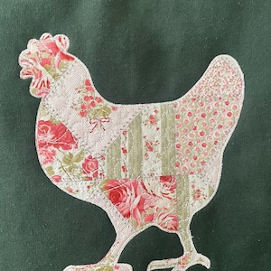 A dark green sweatshirt with an appliqued chicken. The chicken is made of a printed patchwork fabric with pink and green floral patterns. The text 'Weekend Vibes Apparel' is printed in white on a pink background in the lower right corner of the image.
