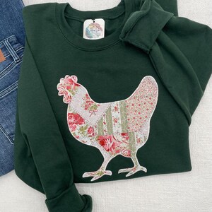 Appliquéd chicken on green sweatshirt. Chicken is appliquéd using a patchwork printed fabric of sage green and pinks on a white background. The appliquéd fabric has a white zigzag thread border and white stippling.