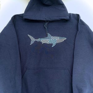 Navy blue hooded sweatshirt with an embroidered shark design. The shark is embroidered with a variety of patterns in thread shades of blues, white, and gray. The sweatshirt has a front kangaroo pocket and a drawstring hood.