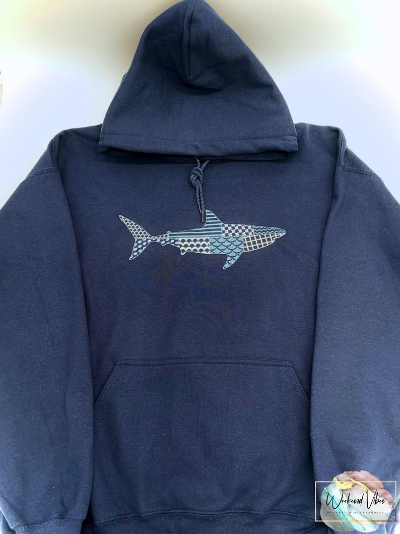Navy blue hooded sweatshirt with an embroidered shark design. The shark is embroidered with a variety of patterns in thread shades of blues, white, and gray. The sweatshirt has a front kangaroo pocket and a drawstring hood.