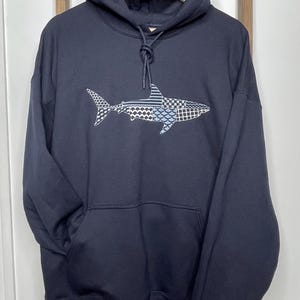 May include: Navy blue hooded sweatshirt with a shark graphic. The shark design features a mix of patterns in white and blue. The sweatshirt has a front pocket and a drawstring hood. The brand name "Weekend Vibes" is visible in the upper right corner.