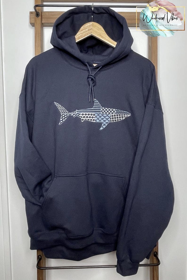 Navy blue hooded sweatshirt with an embroidered shark design. The shark is embroidered with a variety of patterns in thread shades of blue, white, and gray. The sweatshirt has a front kangaroo pocket and a drawstring hood. The brand name "Weekend Vibes" is visible in the upper right corner.