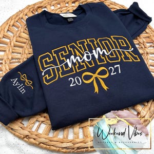 May include: A navy blue crewneck sweatshirt featuring the gold embroidered text SENIOR with the white cursive word mom overlaid. Below, the year 2027 is embroidered in gold with a gold bow icon. The sleeve features the name Aylin and a gold bow.