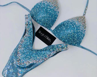 May include: A light blue bikini set with sparkling embellishments. The top and bottom are covered in iridescent sequins, with a gradient effect from light blue to a lighter, almost white color. The set includes a triangle top and a high-cut bottom. The label reads "Master Bikinis".