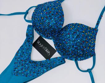 May include: A turquoise bikini set with sparkling blue and multicolored embellishments. The top and bottom are adorned with numerous small, shiny accents. The set includes a triangle top and a thong-style bottom. The tag reads "Master Bikinis".