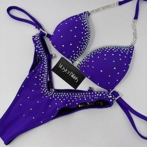 May include: A two-piece purple bikini set with rhinestone embellishments. The top features rhinestone-studded cups and straps, while the bottom has rhinestone accents. The set includes a tag that reads "Master Bikinis".