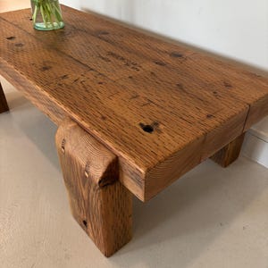 May include: A rustic, rectangular wooden coffee table with a natural finish. The table features thick, solid legs and a tabletop with visible wood grain and markings. A vase of orange flowers sits on the table.