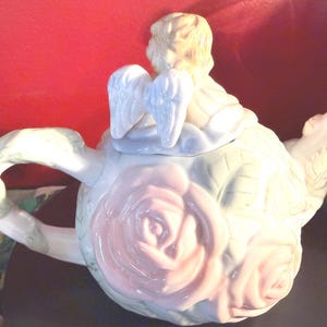 May include: A decorative teapot with a cherub figurine on the lid. The teapot is white with pink roses and green leaves. The handle and spout are also white with green accents. The cherub has light blonde hair and blue-tinted wings.