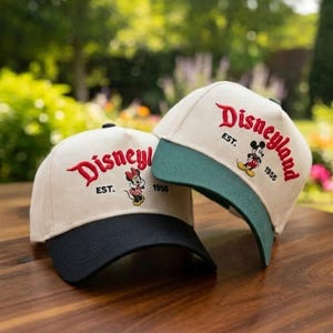 May include: Two baseball caps with the Disneyland logo embroidered in red. One cap has a black brim, the other has a green brim. Both caps are beige with the text "Disneyland EST. 1955" and a cartoon character.