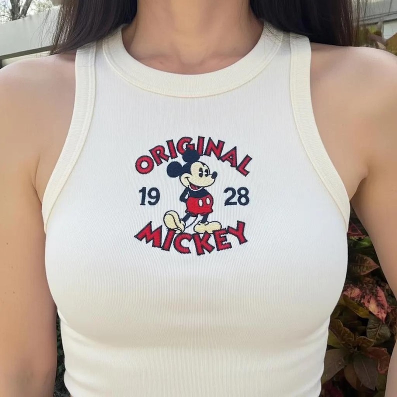 May include: Cream ribbed tank top with a Mickey Mouse graphic. The design includes the word "ORIGINAL" above the cartoon character, and "19 28 MICKEY" below, in red and black. The top has a rounded neckline and is sleeveless.