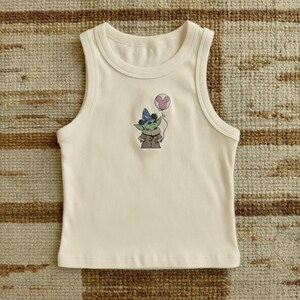 Puede incluir: Camiseta sin mangas color crema con cuello redondo. La parte delantera presenta un personaje de dibujos animados bordado que lleva un gorro de fiesta y sostiene un globo rosa. El personaje es verde, lleva un abrigo marr&oacute;n y tiene ojos negros.