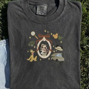 May include: Dark grey Comfort Colors t-shirt with an embroidered design. The design features a cartoon character in a frame, binoculars, a magnifying glass, a car, and a cartoon dog. The shirt is laid on a grassy surface.