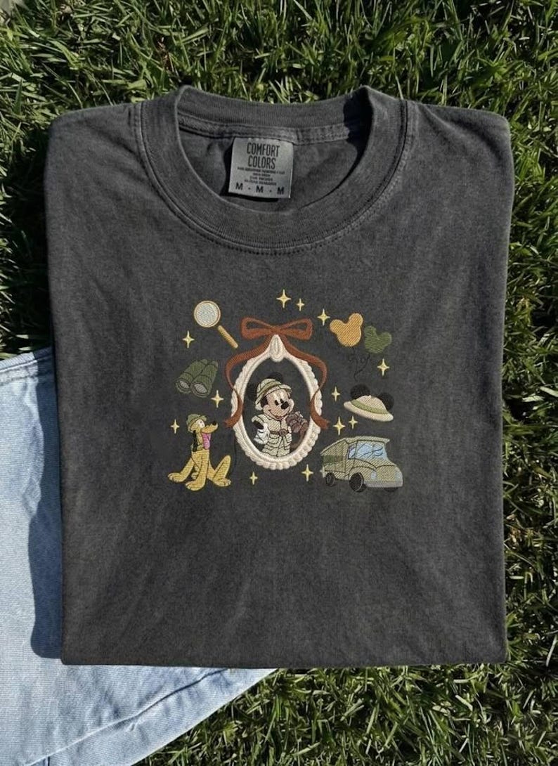 May include: Dark grey Comfort Colors t-shirt with an embroidered design. The design features a cartoon character in a frame, binoculars, a magnifying glass, a car, and a cartoon dog. The shirt is laid on a grassy surface.