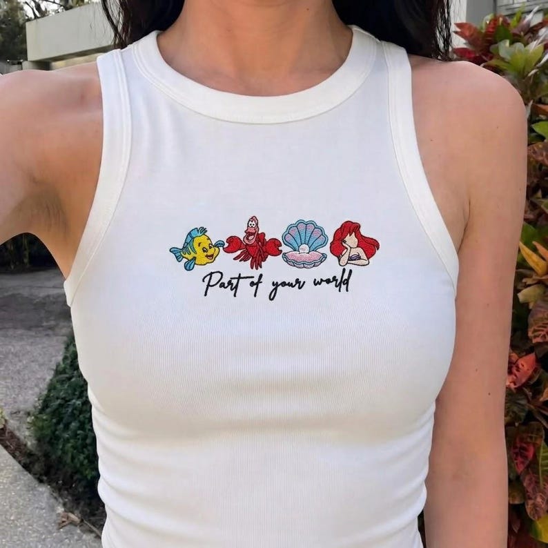 May include: White tank top with a crew neck featuring embroidered cartoon characters and the text "Part of your world" in black script. The design includes a yellow fish, red crab, seashell, and red-haired mermaid.