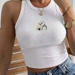 May include: White ribbed tank top with a small embroidered design of two figures embracing. The sleeveless top has a round neckline and is made of a soft, ribbed fabric. The embroidered design features a heart above the figures. The top is paired with blue denim jeans.