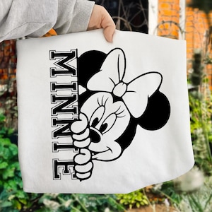 May include: White tote bag featuring a black and white Minnie Mouse graphic. The design includes Minnie's face and bow, with the name "MINNIE" printed vertically in a bold font. The bag is held by a person.