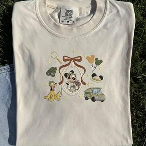 May include: Cream-coloured t-shirt with an embroidered design of Mickey Mouse in safari attire, Pluto, a jeep, binoculars, and a magnifying glass. The shirt has a crew neck and short sleeves. The brand tag is visible at the top.