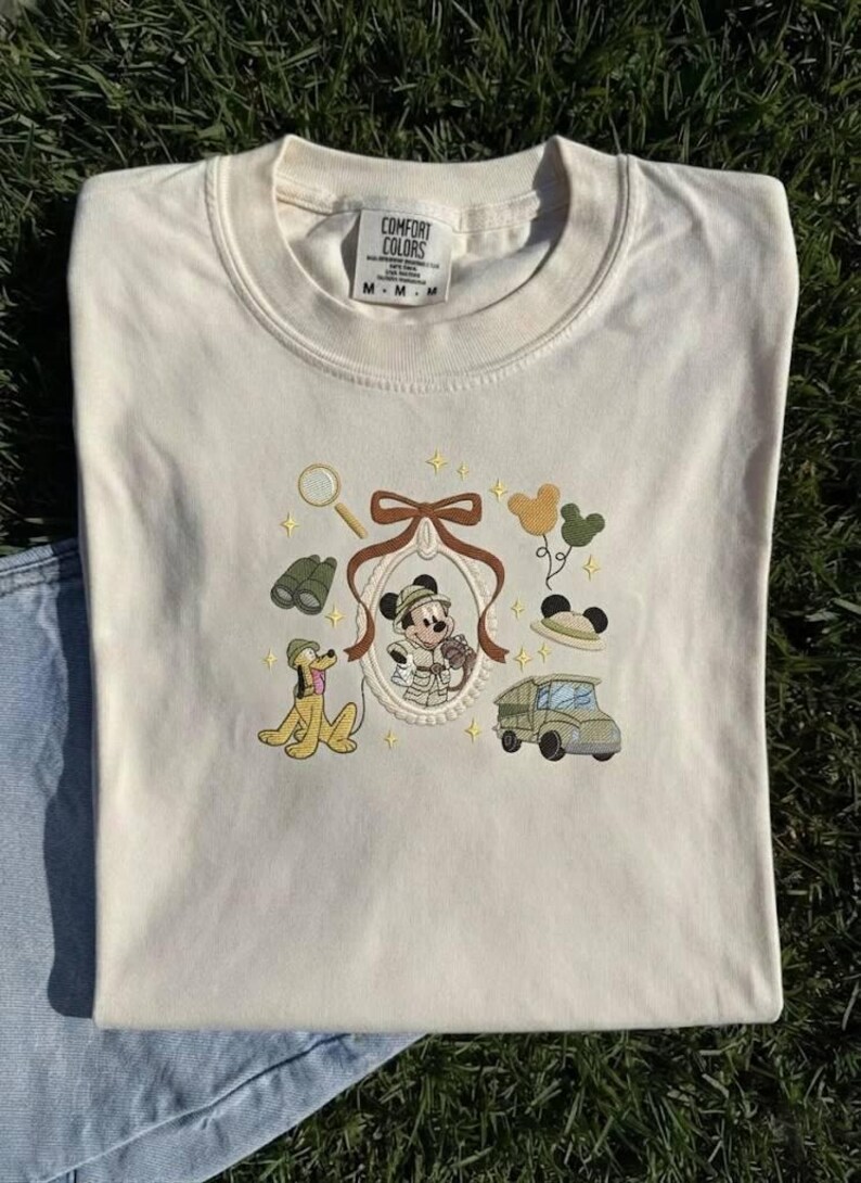 May include: Cream-coloured t-shirt with an embroidered design of Mickey Mouse in safari attire, Pluto, a jeep, binoculars, and a magnifying glass. The shirt has a crew neck and short sleeves. The brand tag is visible at the top.