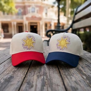May include: Two baseball caps with a beige crown and a red and navy blue brim. Each cap features an embroidered gold sun design with purple floral accents. The caps are displayed on a wooden surface.