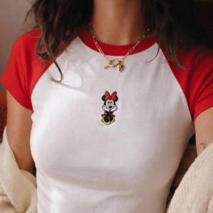 May include: White t-shirt with red sleeves featuring an embroidered Minnie Mouse design. The shirt has a classic baseball tee style. A gold necklace with a heart pendant is also visible.