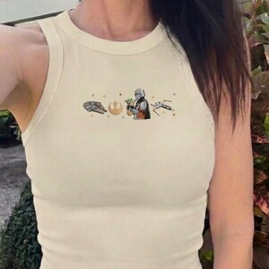 Puede incluir: Camiseta sin mangas color crema con un gr&aacute;fico tem&aacute;tico de Star Wars. El gr&aacute;fico presenta el Halc&oacute;n Milenario, un s&iacute;mbolo de la Alianza Rebelde, el Mandaloriano con Grogu y un caza X-wing. La camiseta es sin mangas y tiene cuello redondo.
