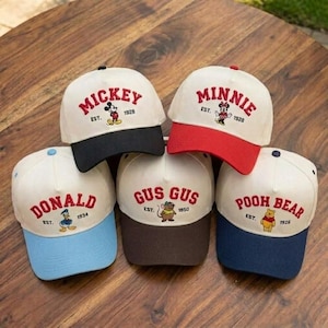 May include: Five baseball caps with white crowns and colored brims. Each cap features a different Disney character's name embroidered in red, along with the character's image and the year of establishment. The brims are black, red, blue, and brown.