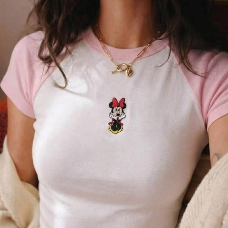 May include: White t-shirt with pink sleeves featuring a Minnie Mouse patch. The shirt has a classic baseball tee design. A gold necklace with a heart pendant is also visible.