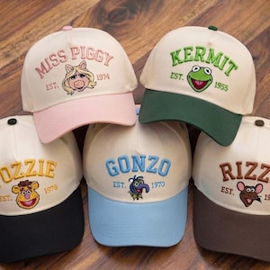 May include: Five baseball caps featuring embroidered Muppet characters. Each cap has a cream crown and a coloured brim. The caps display the names Miss Piggy, Kermit, Fozzie, Gonzo, and Rizzo, along with their respective establishment years.