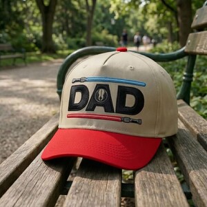 May include: A tan baseball cap with a red brim and top button. The word "DAD" is embroidered in black, with blue and red lightsaber designs above and below the letters. The cap is resting on a wooden bench in a park.