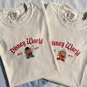 Può includere: Due magliette color crema con la scritta "Disney World" ricamata in rosso. Ogni maglietta presenta un personaggio di Mr. Potato e l'anno "1971". Le magliette sono in materiale morbido e hanno un classico girocollo.