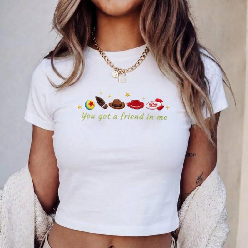 May include: A white short-sleeved t-shirt featuring a graphic print of a colorful ball, a footprint, a brown cowboy hat, a red hat, and a red rocket ship. Below the icons, the text reads "You got a friend in me" in a light green cursive font.
