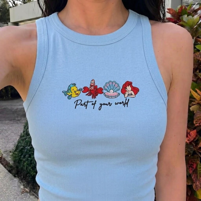 May include: Light blue ribbed tank top with a graphic of cartoon characters and the text "Part of your world". The graphic includes a yellow fish, a red crab, a seashell, and a red-haired mermaid.