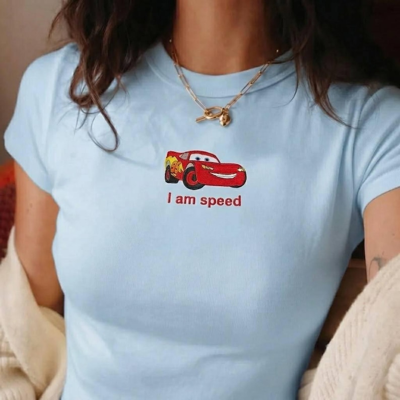 K&ouml;nnte beinhalten: Hellblaues T-Shirt mit einem roten Cartoon-Rennwagen und dem roten Text "I am speed". Das T-Shirt hat kurze &Auml;rmel und einen Rundhalsausschnitt.